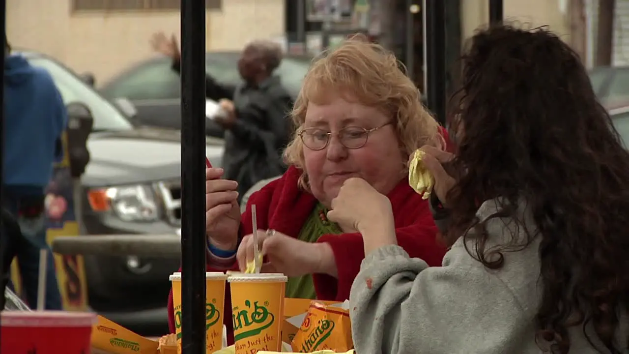 Two Adult Females Eating Fast Food Outside Sitting On Table With People And Traffic Going Past In Background