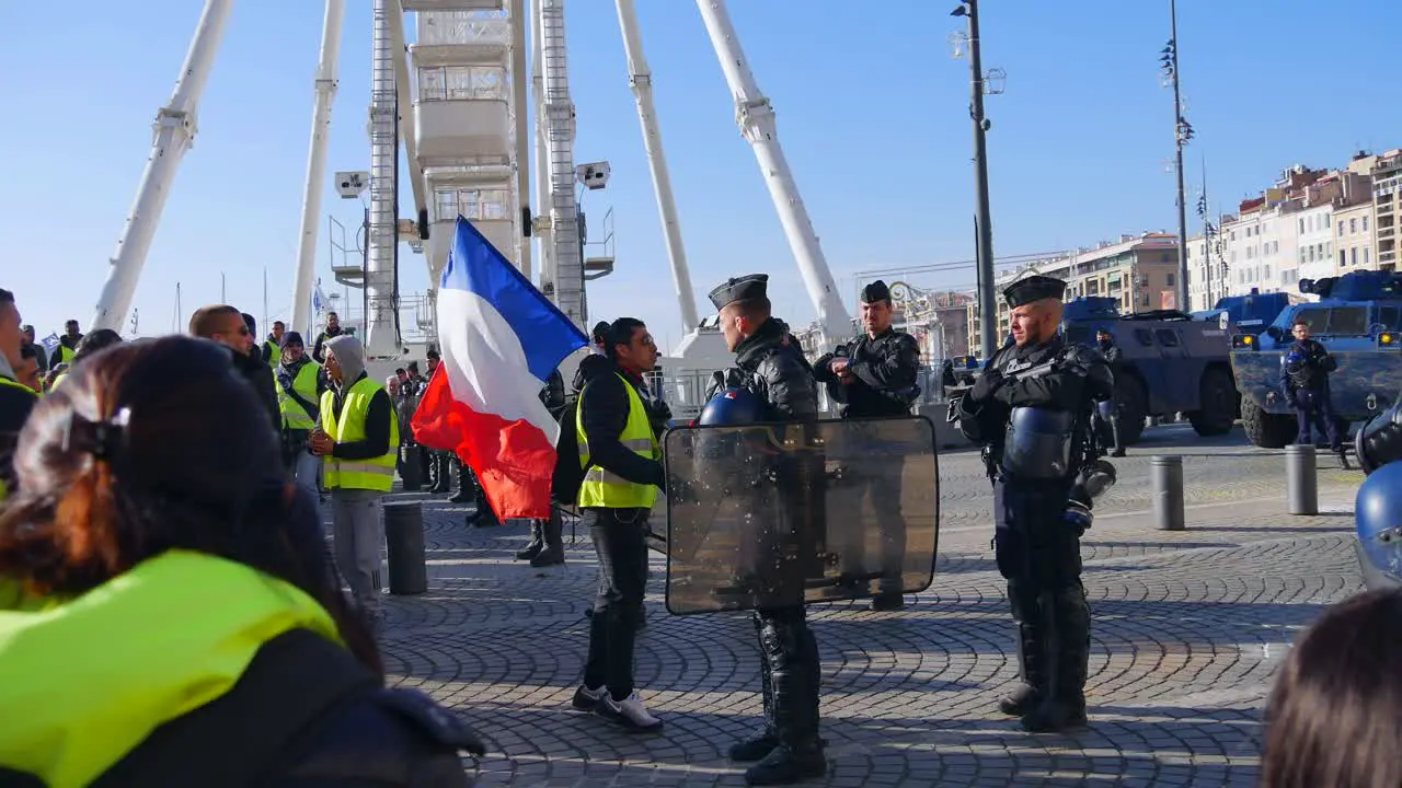 Yellow jacket demonstrator with a french flag and sun glasses walks towards a police officer in full riot gear and points a finger at him