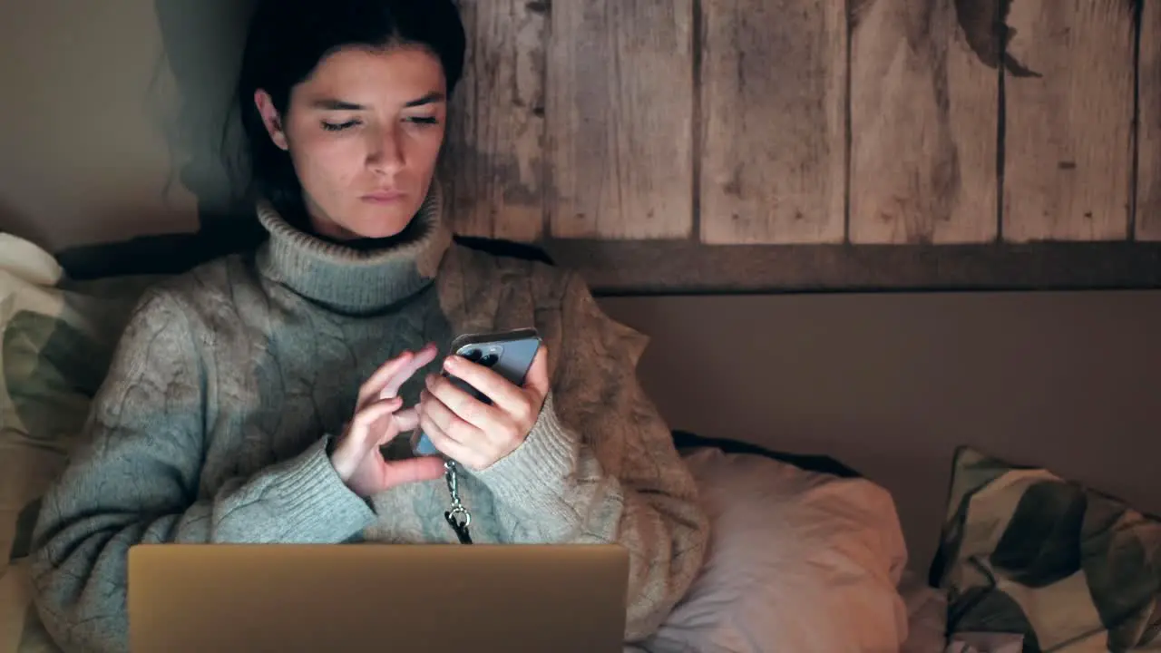 Young Caucasian woman browsing on phone and laptop while lying in bed at home