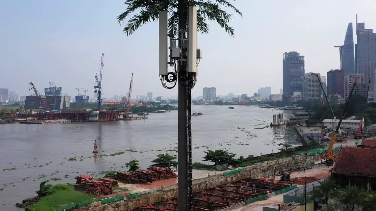 Ascending aerial shot of ultra modern mobile phone tower disguised as a palm tree against a background of a riverfront construction site with city buildings cranes and boat traffic on the river