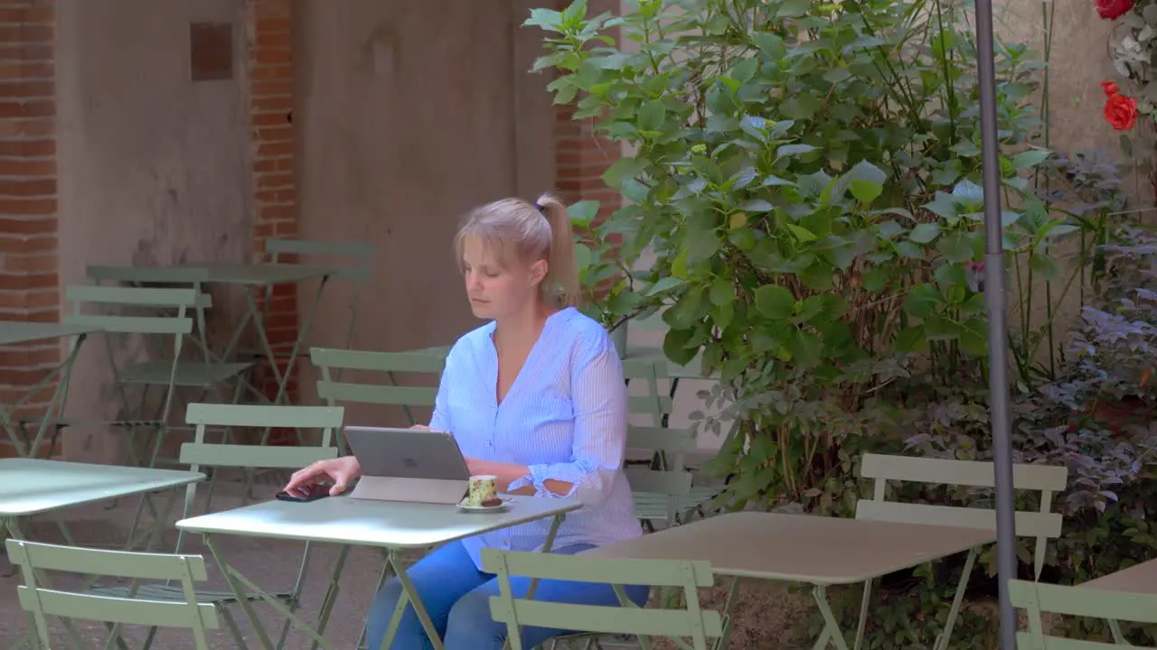 A blonde Caucasian woman with her hair tied back sitting on a café terrace using her tablet smartphone and having her coffee