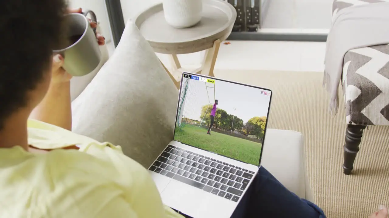 Video of biracial woman sitting on sofa and watching football on laptop at home