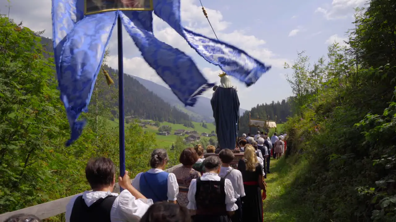 Female worshippers in traditional Tyrolean costumes carry the Virgin mary and a blue banner in a mountain landscape during a religious procession