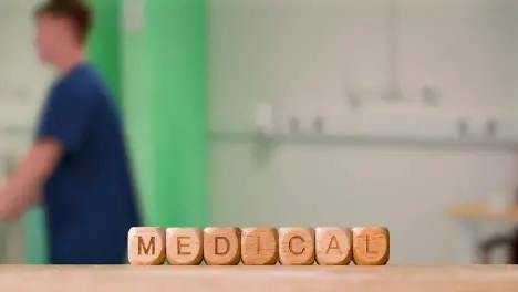 Concept With Wooden Letter Cubes Or Dice Spelling Medical Against Background Of Nurse Wheeling Patient In Hospital Bed