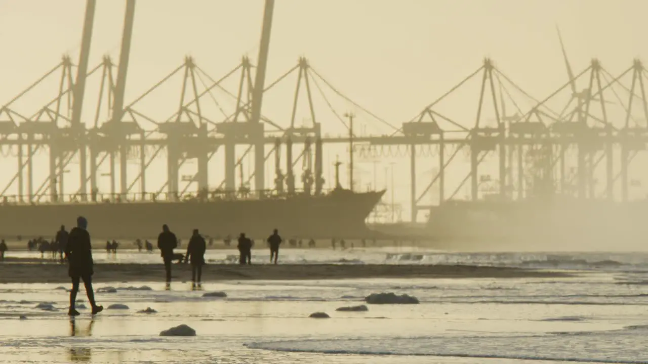 People silhouettes walk on beach in sunset with industrial port crane backdrop