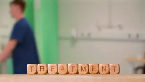 Medical Concept With Wooden Letter Cubes Or Dice Spelling Treatment Against Background Of Nurse Wheeling Patient In Hospital Bed