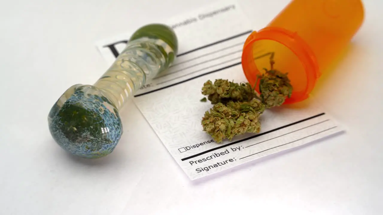 A glass pipe on a white table with marijuana buds spilling out of an orange bottle onto a prescription note