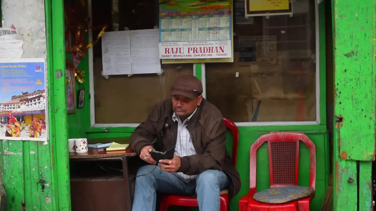 A 40 year old Indian male sitting in one of the shops in the Darjeeling street market and checking his phone