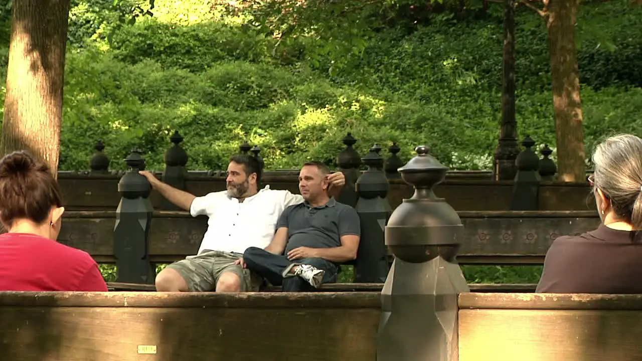 Same Sex Couple Sitting On Bench In Public Park With People Going By