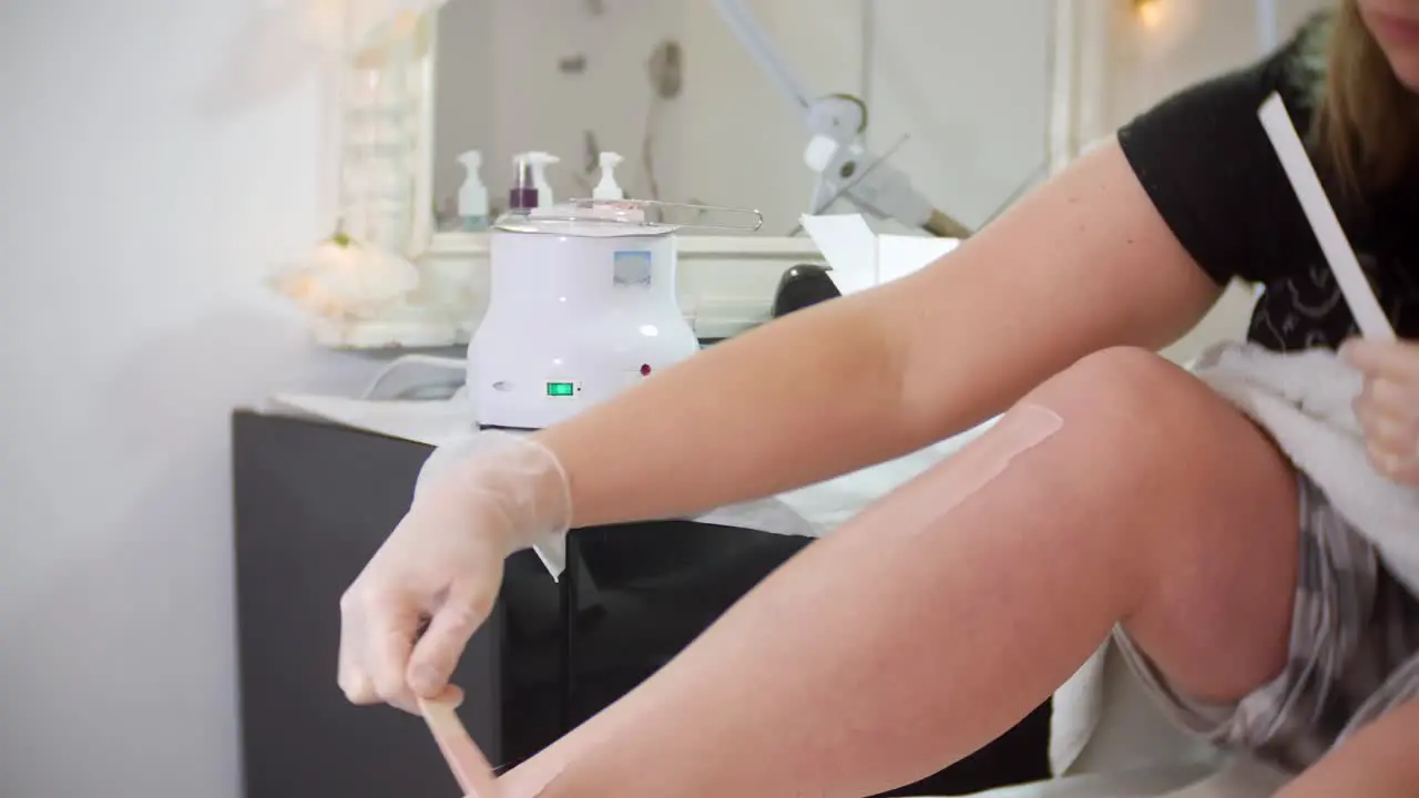 A woman is waxing her own legs using hot wax in a beaty salon treatment room with a spatula