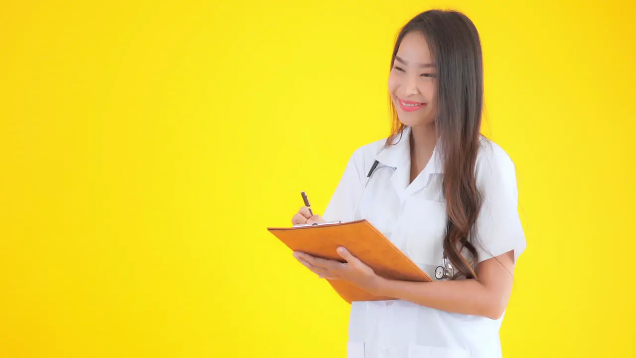 Asian nurse doctor woman writing notes in the clipboard on yellow isolated background smiling