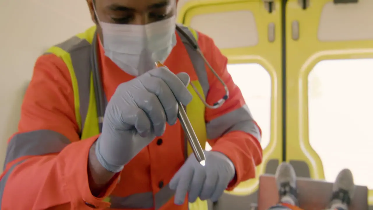 Paramedic Wearing Facial Mask Using A Flashlight And Observing The Eyes Of A Patient Lying On The Stretcher Inside An Ambulance