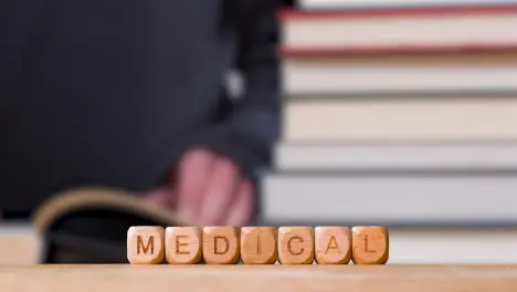 Education Concept Shot With Wooden Letter Cubes Or Dice Spelling Medical With Person Reading Book In Library