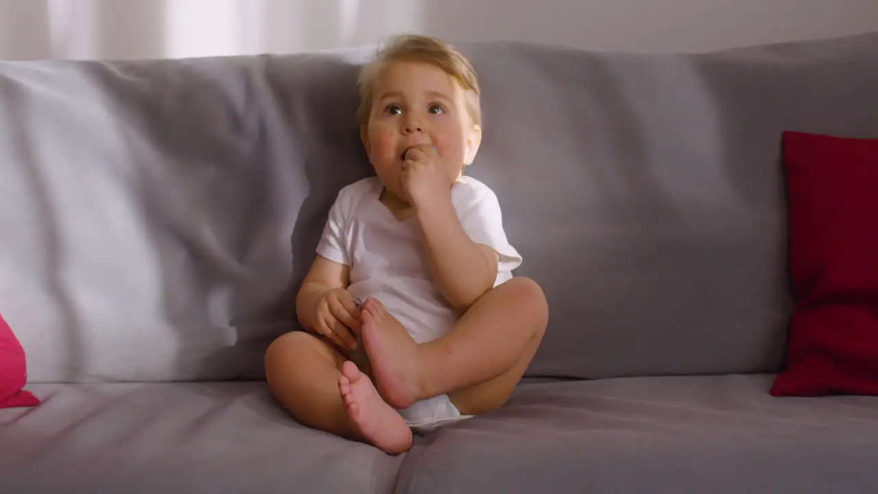 Front View Of A Baby Sitting On Sofa In Living Room At Home While Bitting A Toy Animal