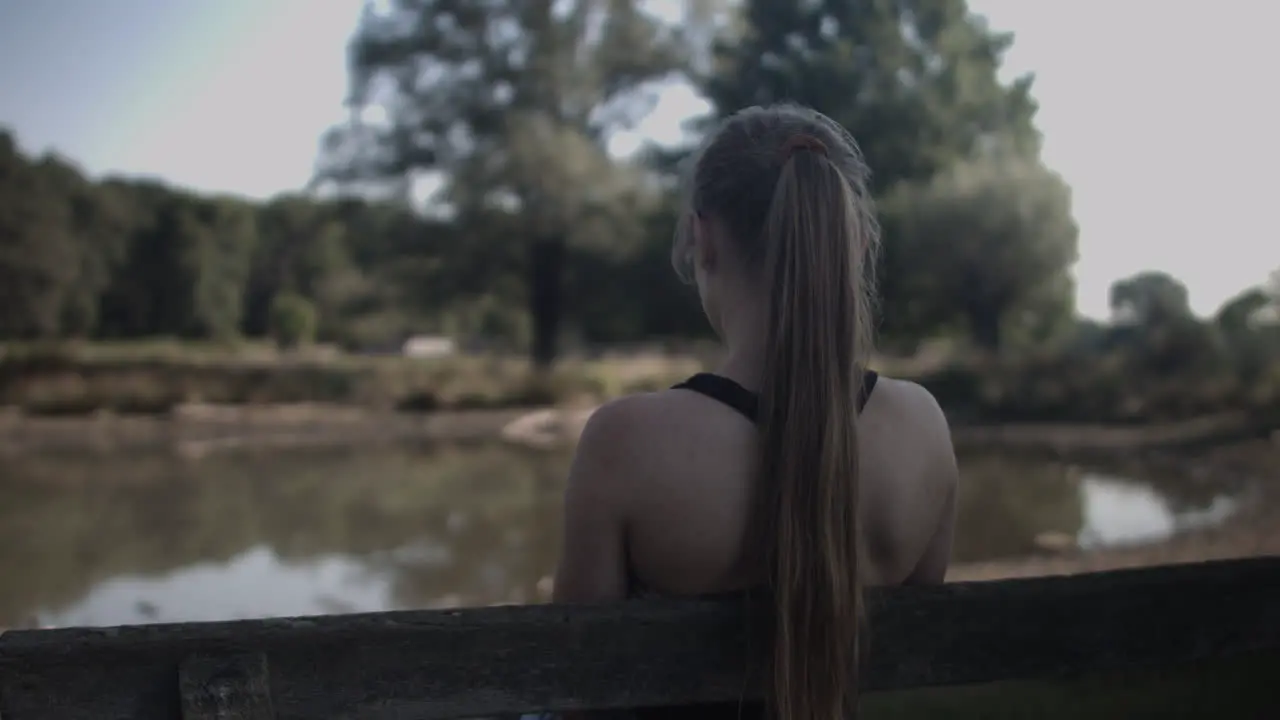 Backside of the young woman sitting under the shade of the tree looking at the still lake in the distance