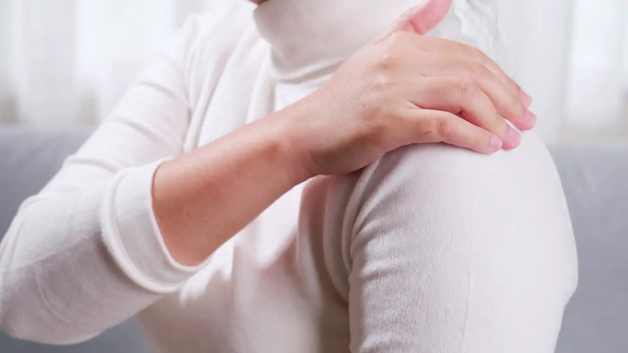 Close up of woman sitting on sofa doing self massage on shoulder and arm for relieving pain and getting relaxation