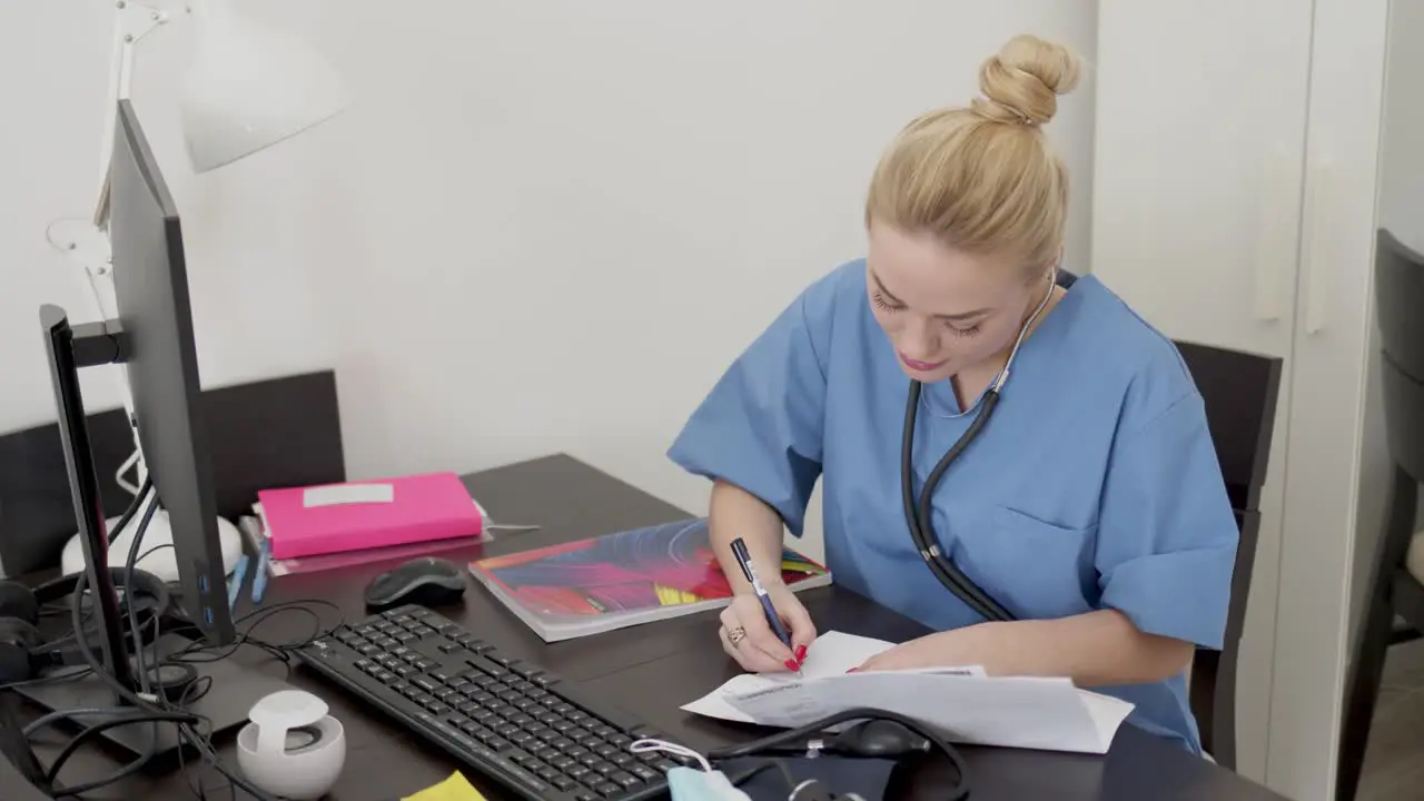 Young beautiful blond female doctor in blue coat working at desk using computer and doing paperwork