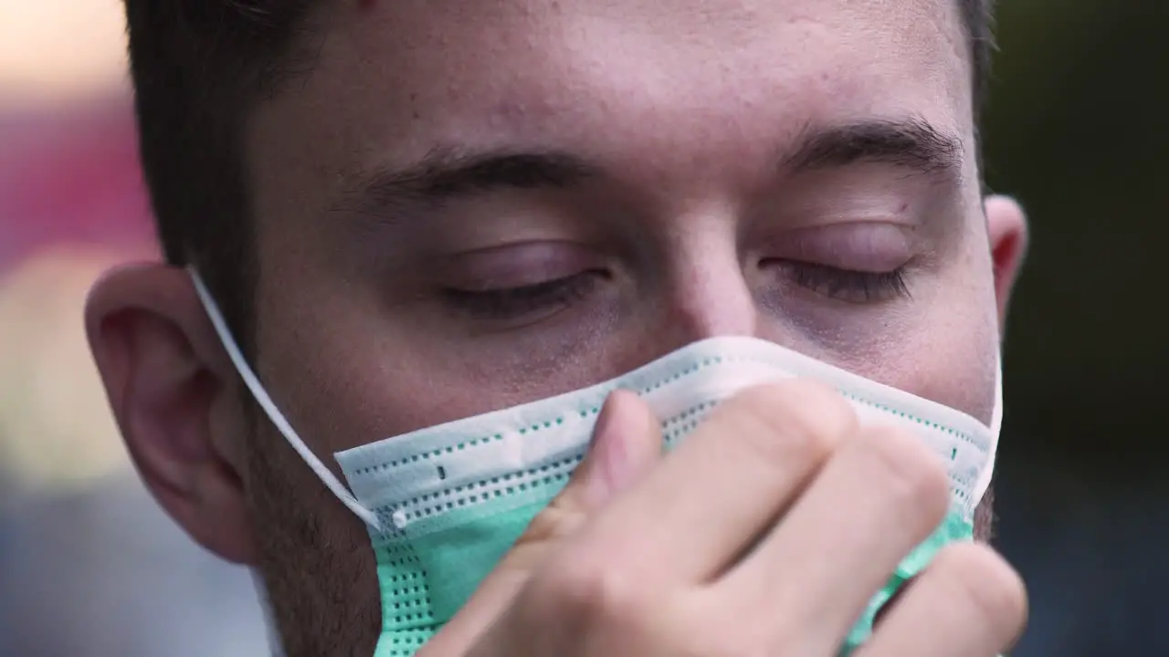 A young man with bright blue eyes and a stubble puts on a green protective Covid-19 facemask in public adjusting it on his nose and face looking straight into the camera static close up 4k shot