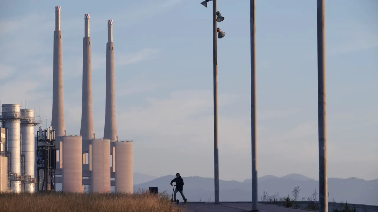 Man contemplating industrial landscape rides off on electric scooter at sunset