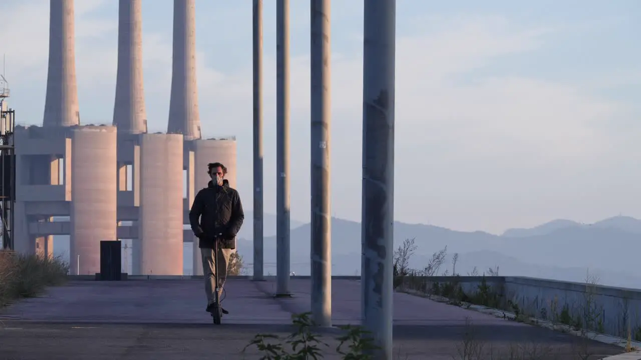 Man with face mask rides electric scooter in sunset industrial landscape