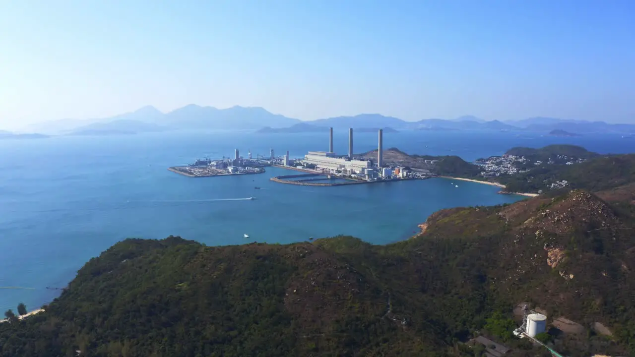 Drone shot traveling to the left over a big green mountain with big coastal coal-fired power plant on an island next to a village with a beach during a beautiful sunny day