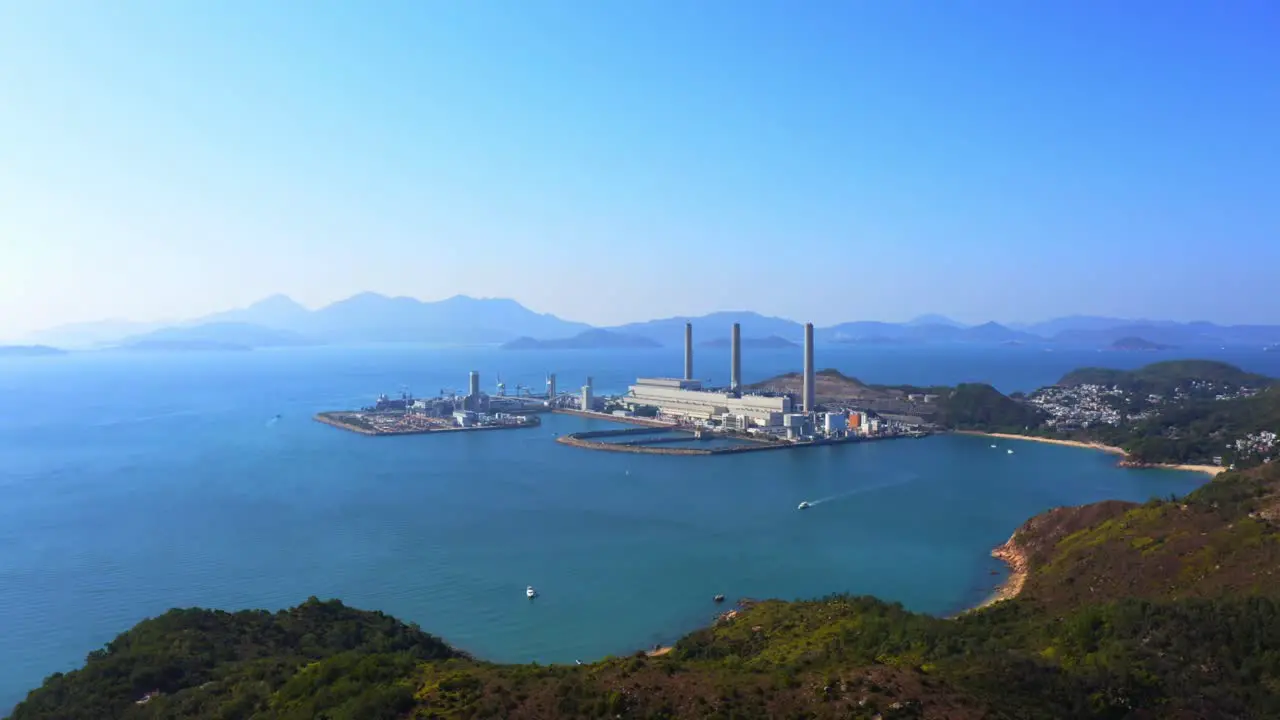 Drone shot traveling backward of a big coastal coal-fired power plant on an island next to a village with a beach during a beautiful sunny day