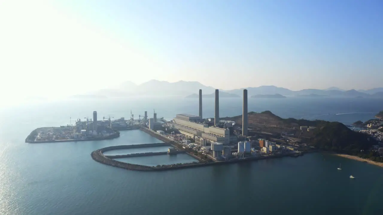 Drone shot traveling to the left of a big coastal coal-fired power plant on an island next to a village with a beach during a beautiful sunny day-2