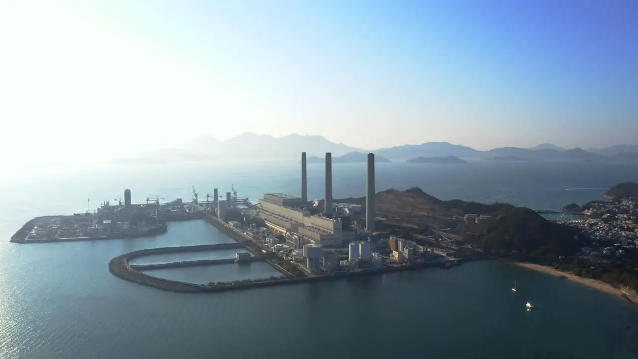 Drone shot traveling to the left of a big coastal coal-fired power plant on an island next to a village with a beach during a beautiful sunny day-1