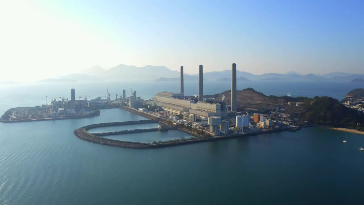 Drone shot traveling to the left of a big coastal coal-fired power plant on an island next to a village with a beach during a beautiful sunny day