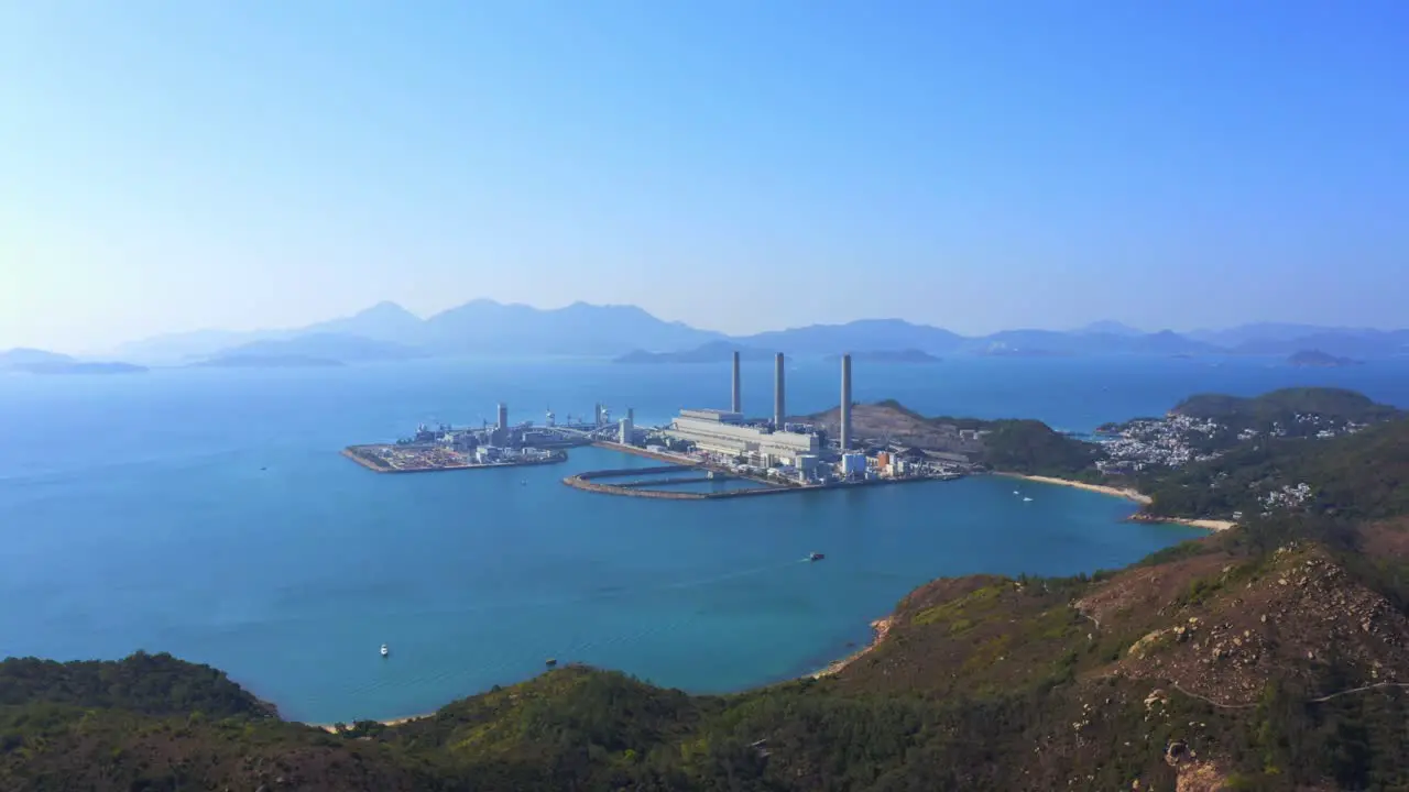 Drone shot traveling backward over a big green mountain with big coastal coal-fired power plant on an island next to a village with a beach during a beautiful sunny day