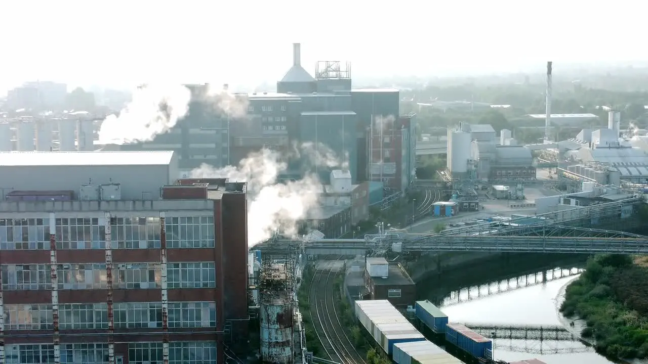 Industrial river chemical warehouse building with factory steam emissions in early morning sunlight aerial descending