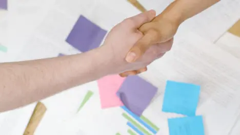 Overhead Close Up Of Business Colleagues Shaking Hands Over Paperwork On Table