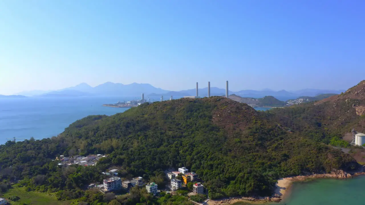 Drone shot traveling forward to a big green mountain and going up to reveal big coastal coal-fired power plant on an island next to a village with a beach during a beautiful sunny day