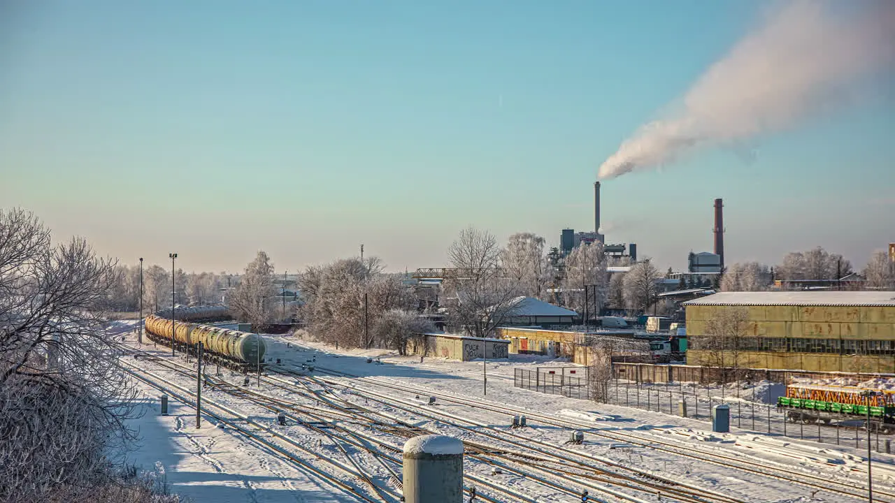 The industrial part of a town in winter with a tanker car train running by as steam come out of a factory smokestack time lapse