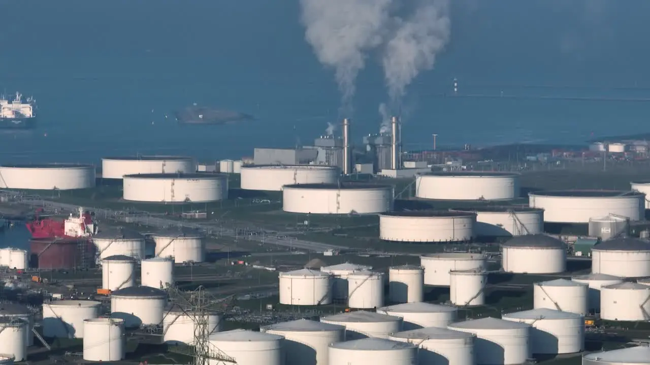 Wide aerial view of large capacity oil storage tanks near the ocean with ships in the background and industrial facility with stacks emitting steam