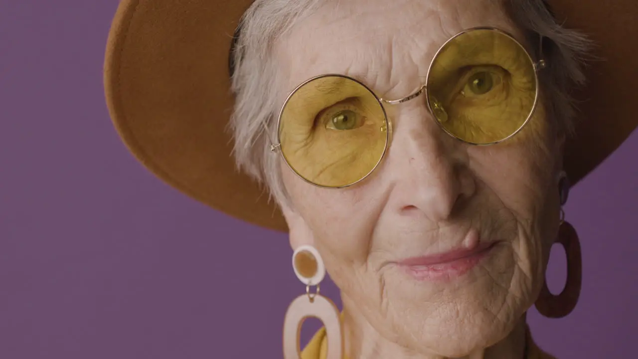 Close Up View Of Senior Woman With Short Hair Wearing Hat Sunglasses And Earrings Posing And Looking At Camera On Purple Background