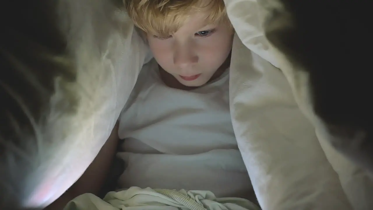 Little Boy Using A Tablet Watching A Movie Covered By The Bed Sheet At Night