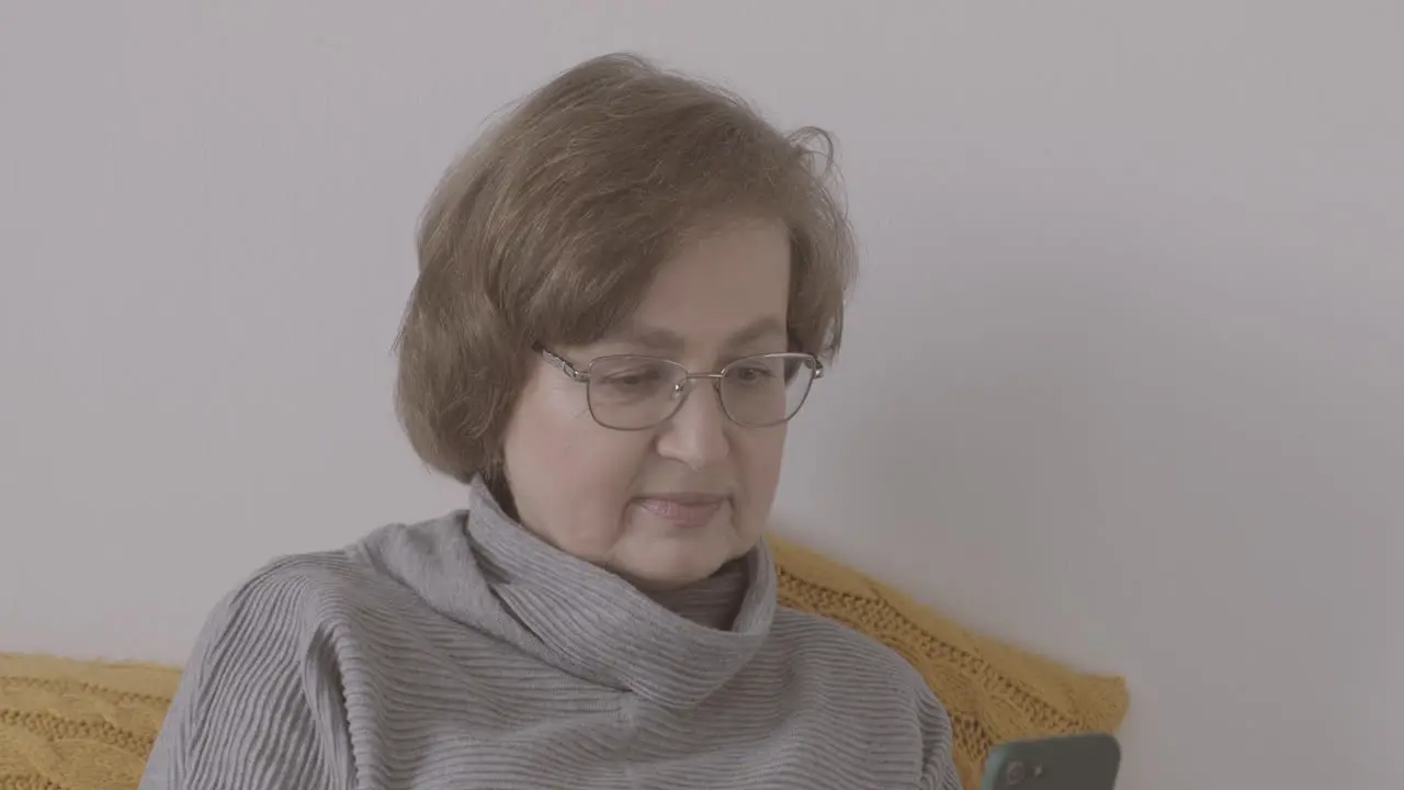 Senior Woman With Eyeglasses Sitting On Bed Using Smartphone 1