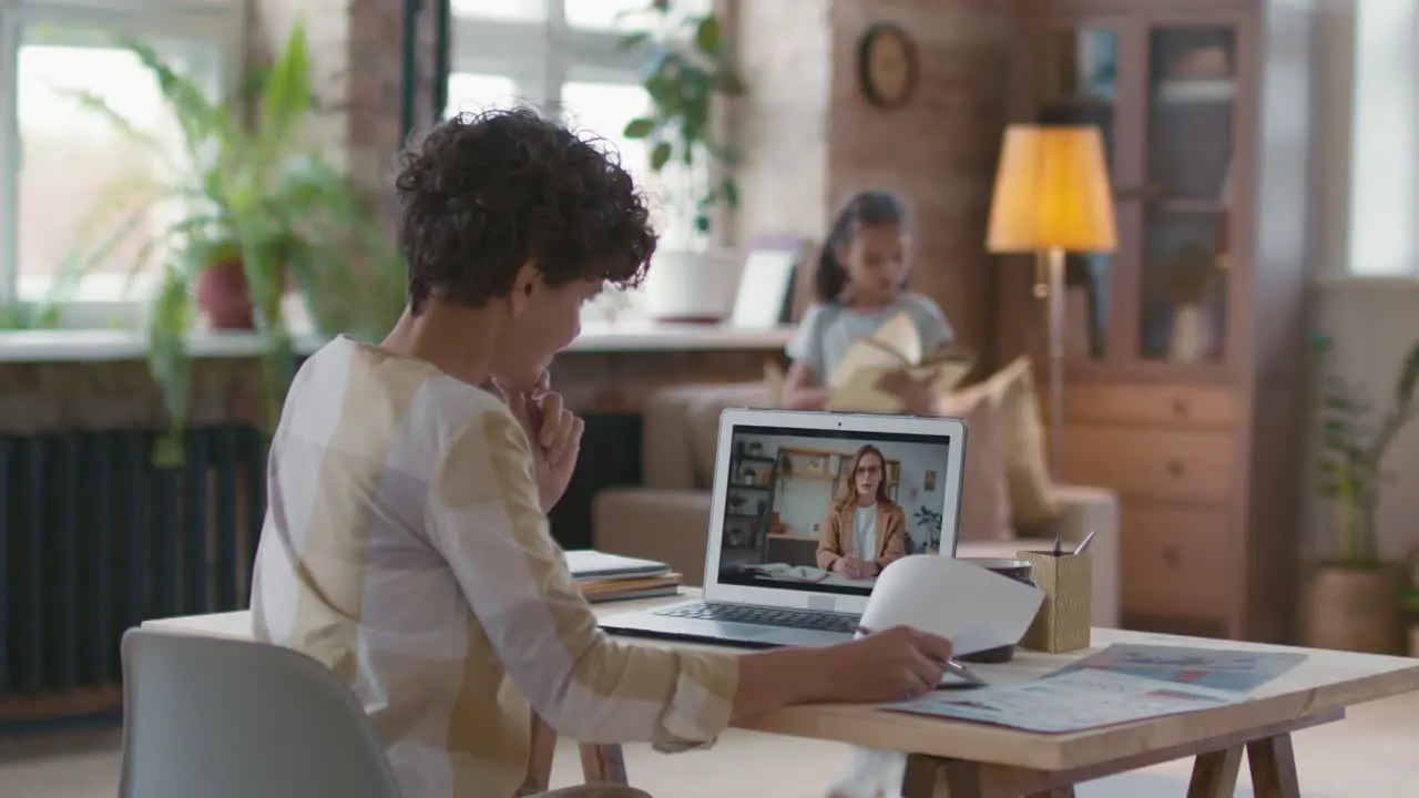 Woman With Short Curly Hair Sitting At A Desk In Front Of The Computer While Presenting A Project In The Background His Daughter Is Reading A Book