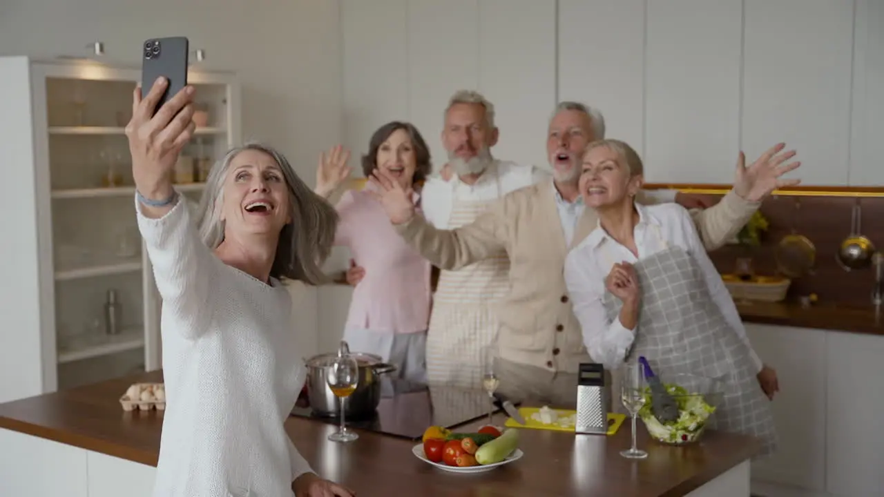 Happy Senior Woman Making A Selfie With A Smartphone In The Kitchen Her Friends Laugh And Wave At The Camera