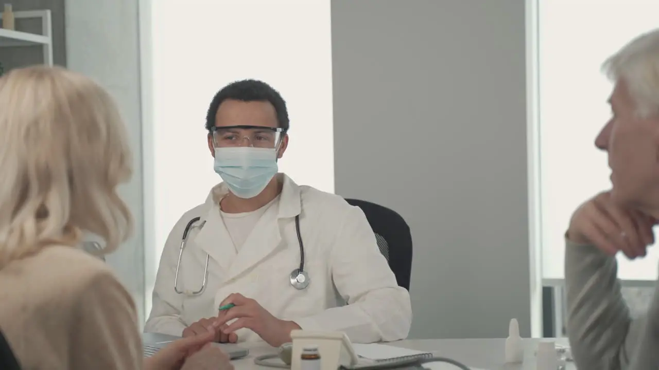 A Young Black Doctor In A Facemask Listening To A Ederly Couple