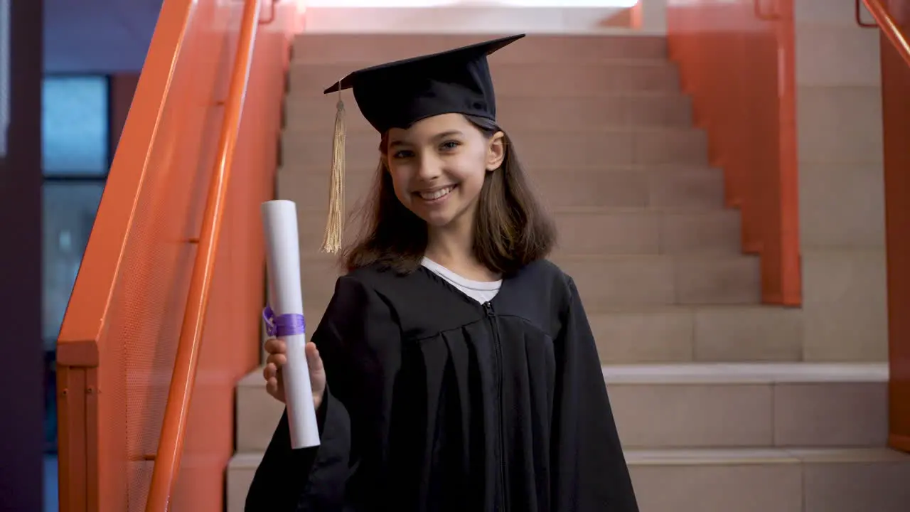 Portrait Of A Happy Preschool Female Student In Cap And Gown Running Down The Stairs Holding Graduation Diploma And Looking At The Camera