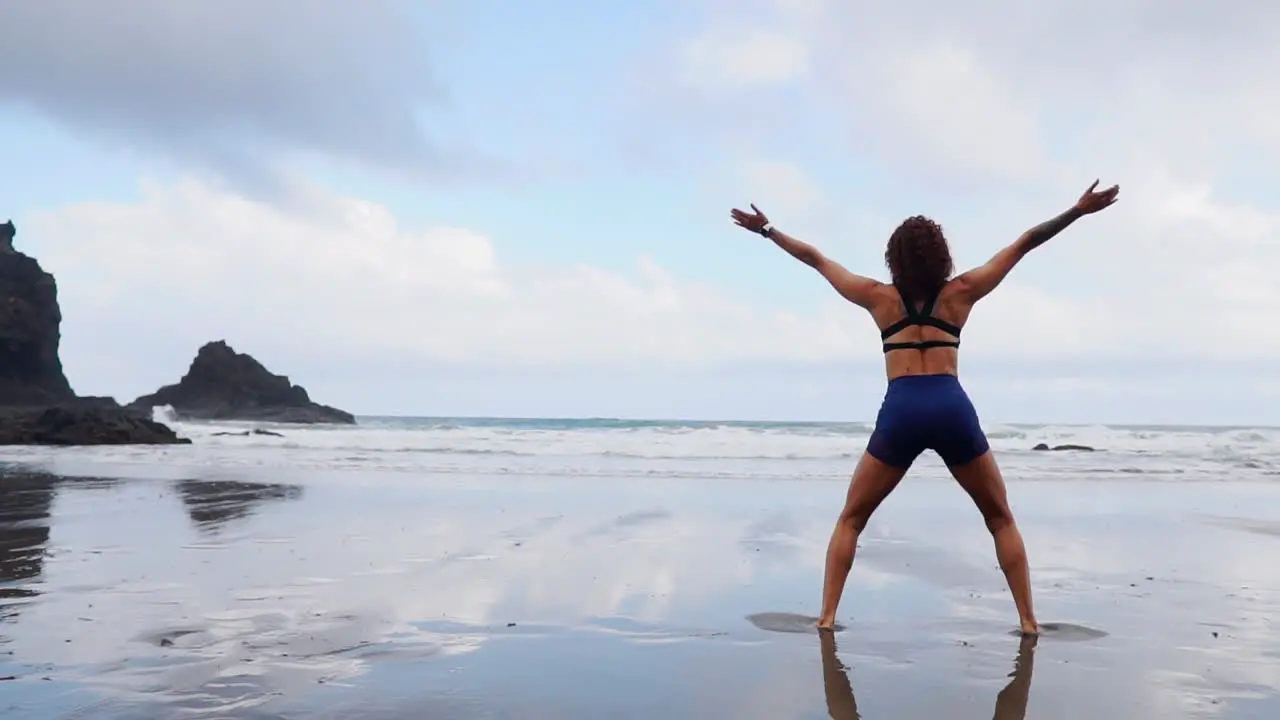 In slow motion a young slim woman practices squats and jumps working out on the ocean's shore