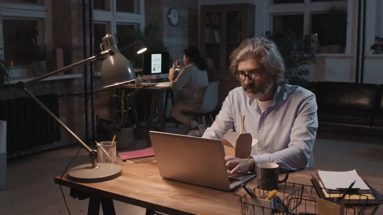 Grey Haired Man With Eyeglasses And Blue Shirt Sitting In Front Of Computer While Eating Food And Working In The Background His Partner Works Sitting At A Desk