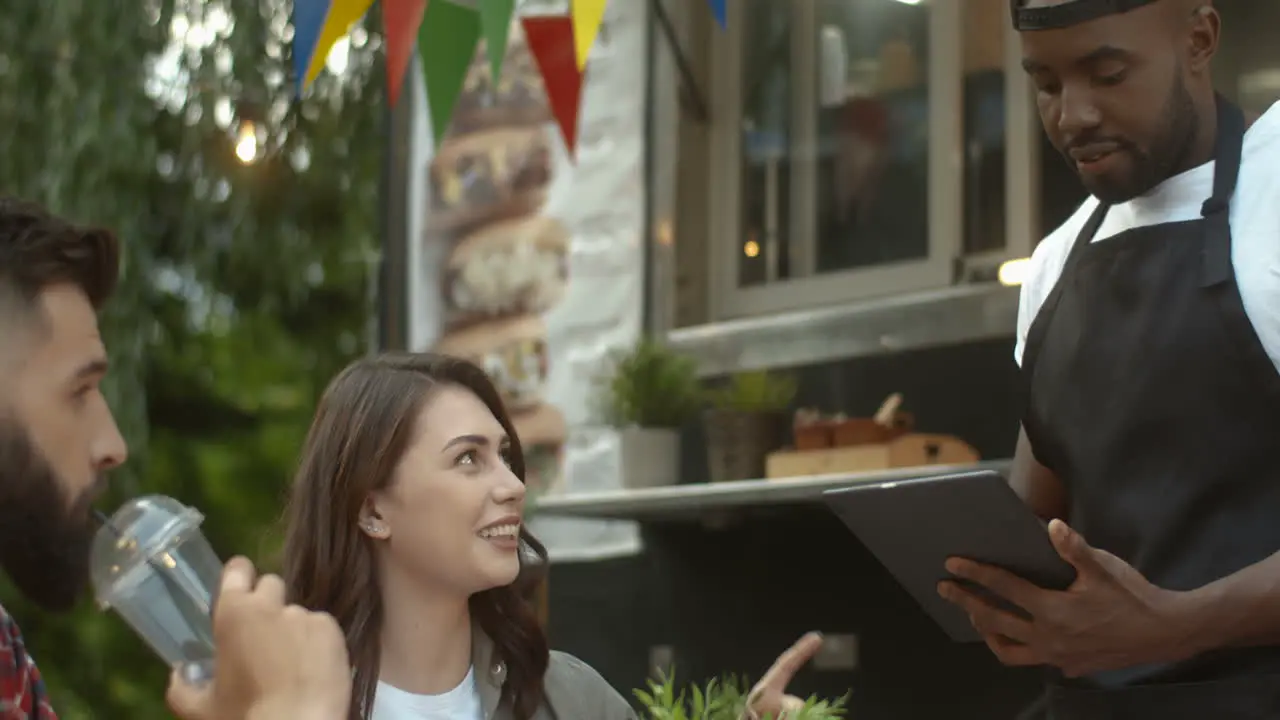 Happy Young Couple Sitting At Table Outdoors In Park At Truck Bar Ordering Food And Drinks From Male Waiter With Tablet