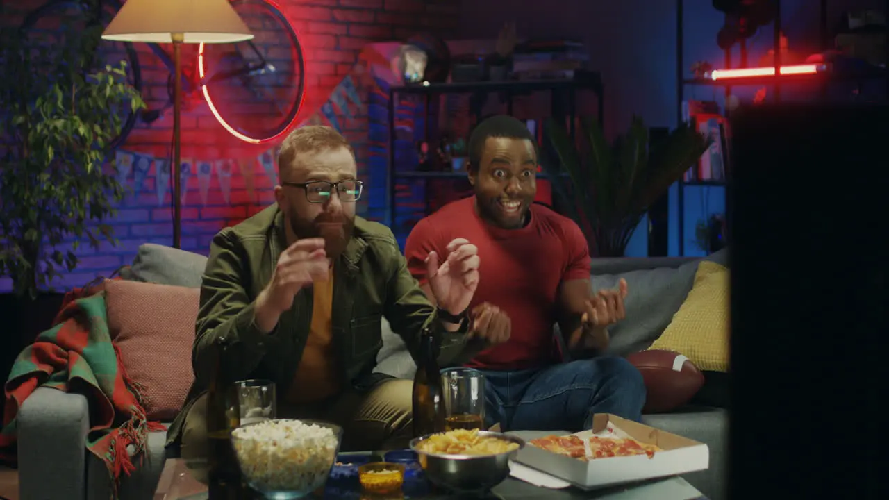 Multiethnic Young Male Sport Fans Sitting On Sofa In The Dark Living Room With Beer And Snacks Watching Sport Game On Tv And Celebrating Victory Of Their Team