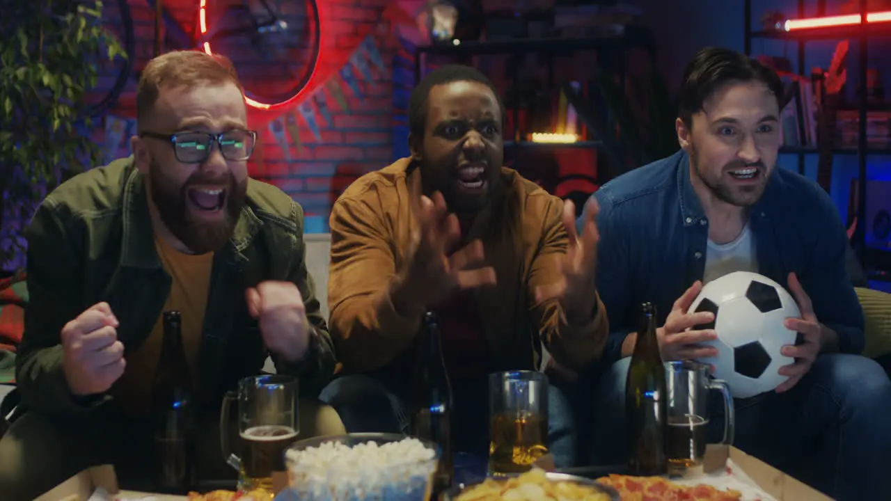 Three Multiethnic Friendly And Cheerful Young Men Seeing Loose Or Missing A Goal Of Their Football Team In The Championship In The Dark Living Room With Snacks And Beer