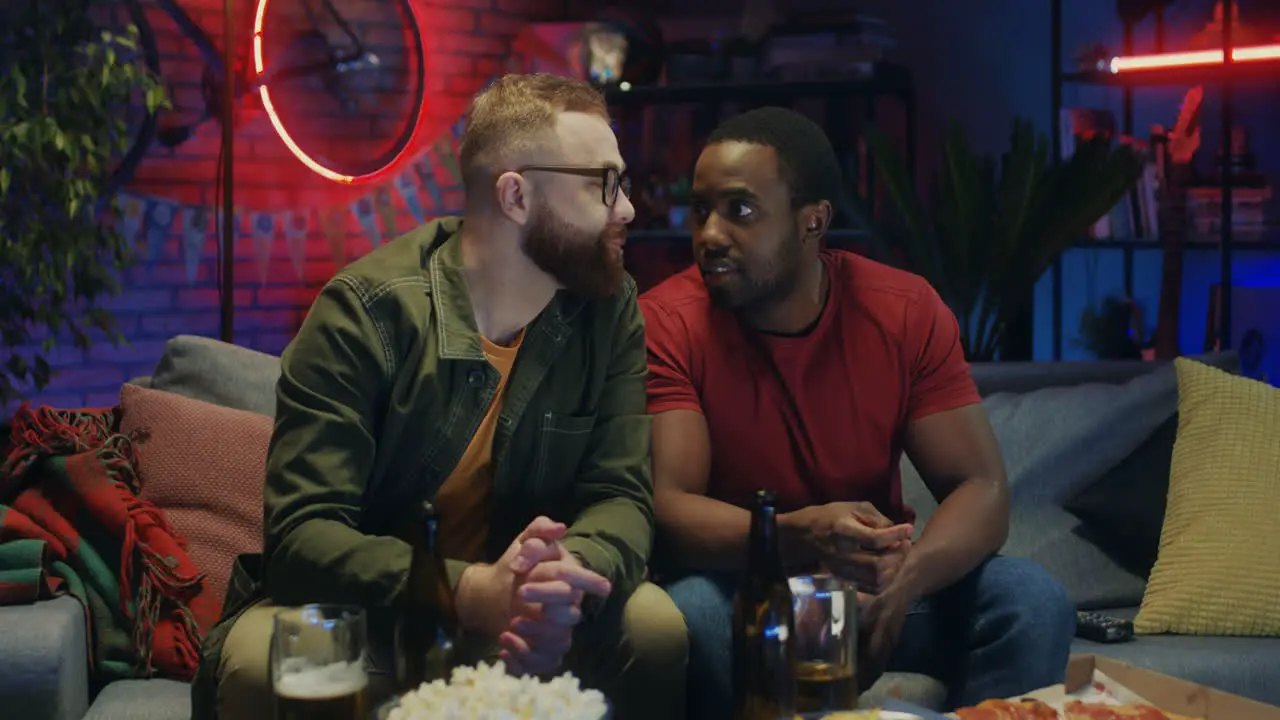 Two Mixed Races Cheerful Guys Sitting On The Sofa With Beer And Snacks Talking And Watching Tv With Sport Channel