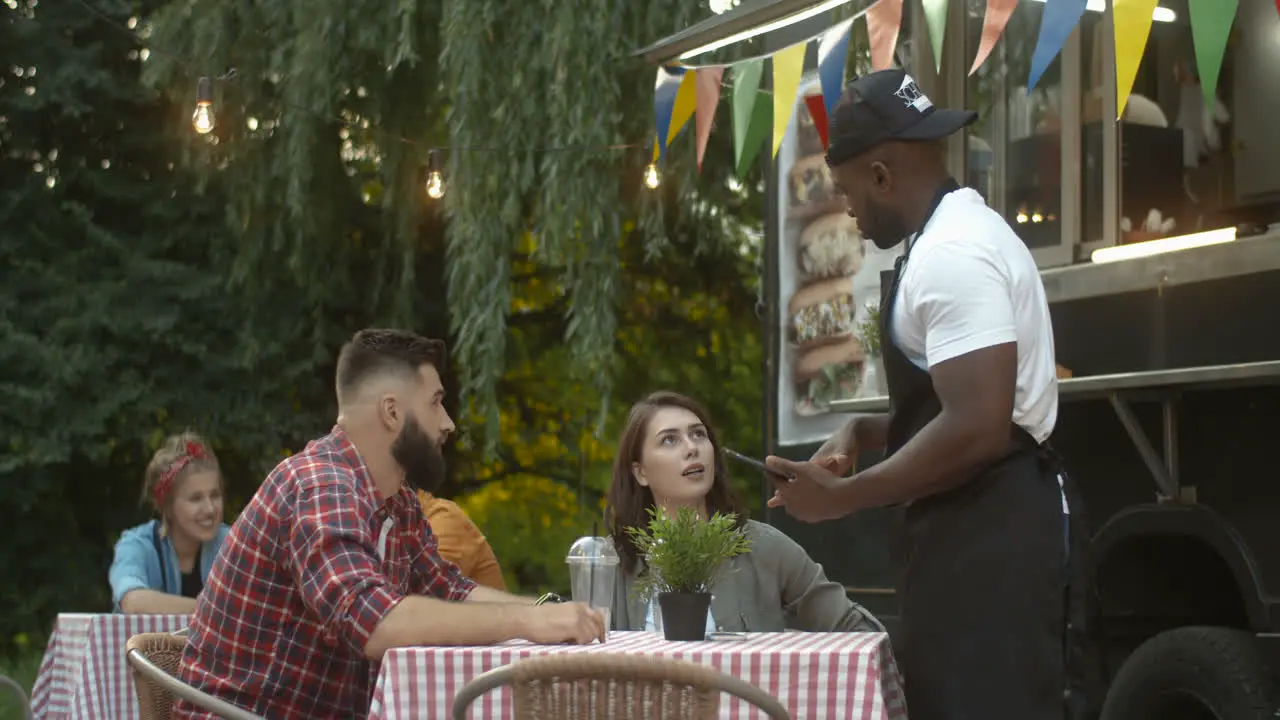 Young Handsome And Cheerful Male Barman In Apron And Cap Asking For Order From Happy Couple At Table Outdoor Small Food Truck Cafe In Park