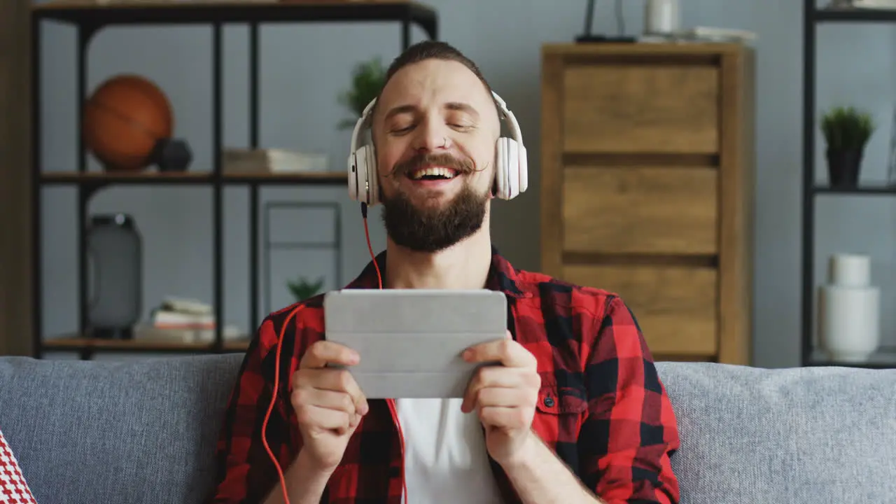 Close Up Of The Handsome Hipster Man In The Big White Headphones Watching Something Funny On The Tablet Computer Nodding His Head And Laughingon The Couch In The Living Room